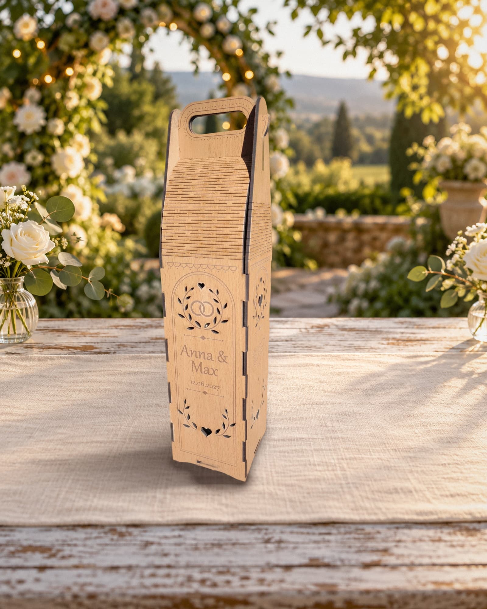 Hochzeits-Weinträger aus Holz mit Beispielgravur Anna & Max und Datum, Lorbeerkranz mit verschlungenen Ringen, auf einem weiß gedeckten Gartentisch vor einem Rosenbogen im Abendlicht